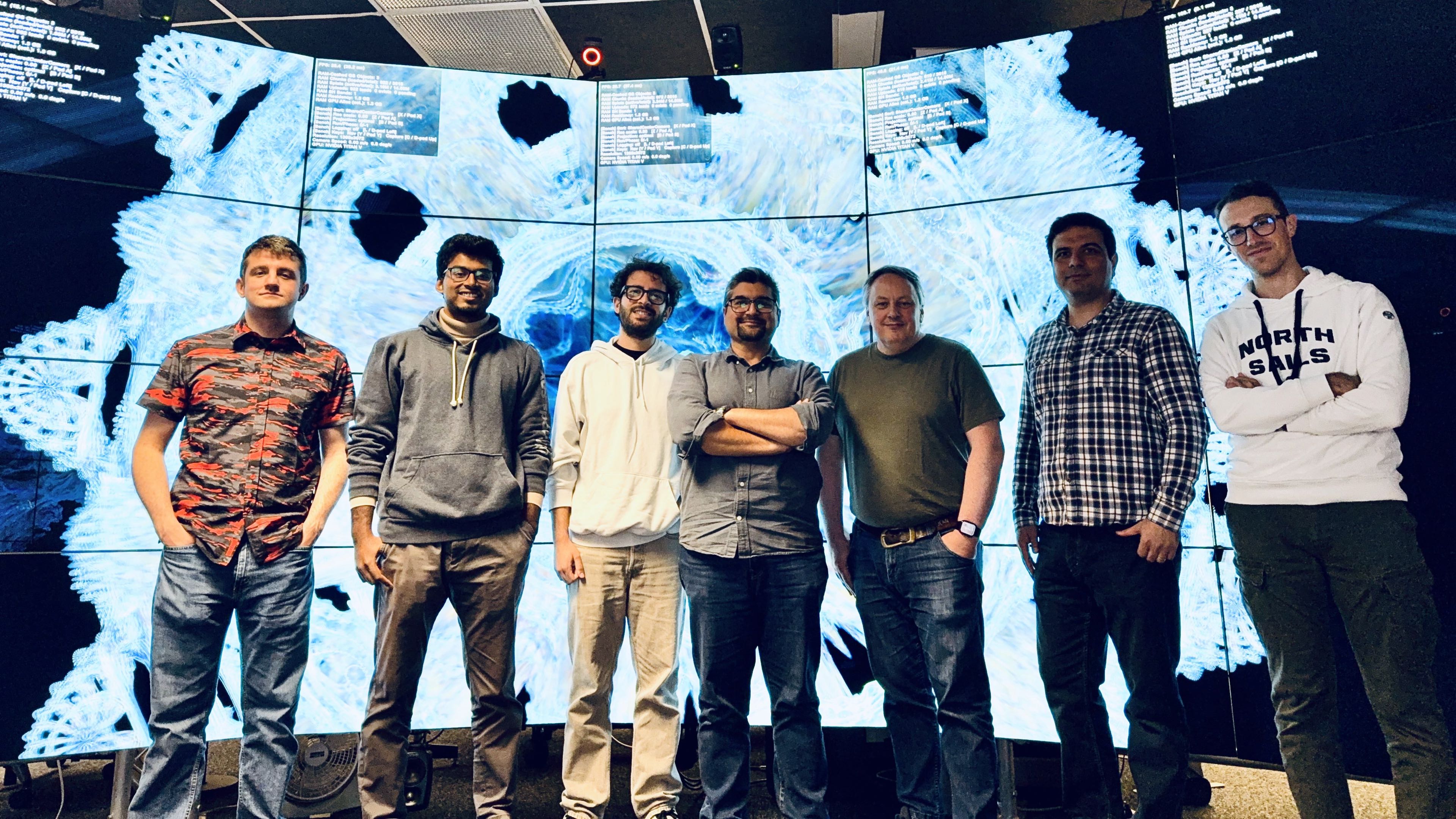 The immersive vis team standing in front of EVL's tiled display wall showing a large blue volumetric visualization